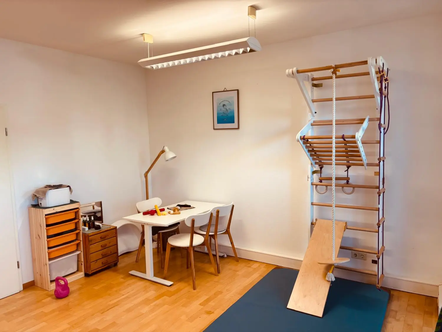 Sentio – ein weißer Sitz-Steh-Schreibtisch mit zwei Stühlen, ein hölzernes Aufbewahrungsmöbel mit orangefarbenen Schubladen und ein hölzernes Klettergerüst mit Rutsche und Ringen auf der rechten Seite